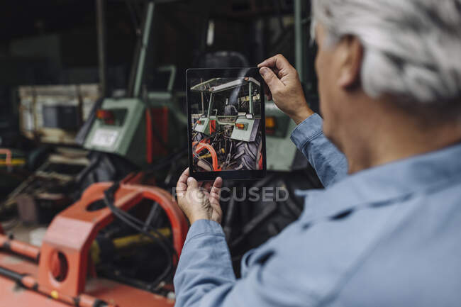 Старший фотографирует трактор в сарае с планшетом — стоковое фото