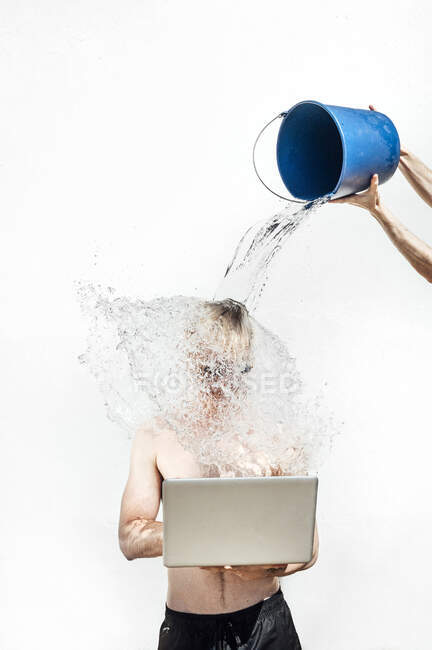 Hands of woman pouring water with bucket on shirtless young man using laptop against wall — Stock Photo