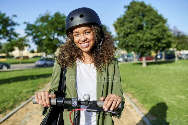 Mujer joven feliz de pie con scooter eléctrico durante el día soleado - foto de stock