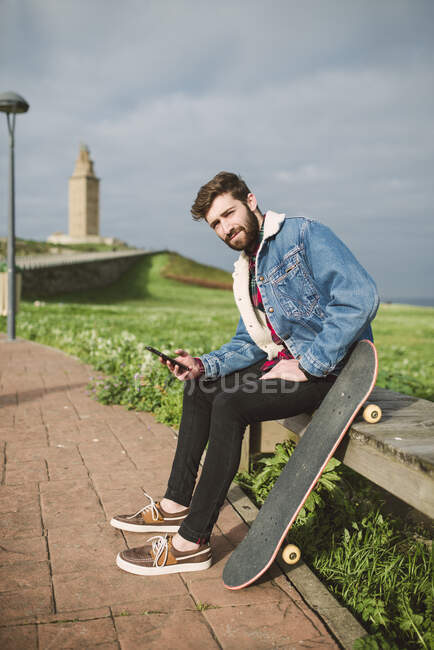 Bello giovanotto che usa lo smart phone mentre è seduto con lo skateboard sul sedile — Foto stock