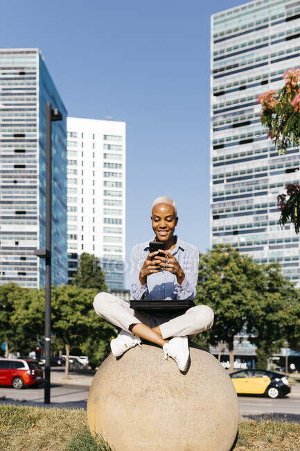 Donna d'affari sorridente utilizzando il telefono mentre seduto sulla roccia in città — Foto stock