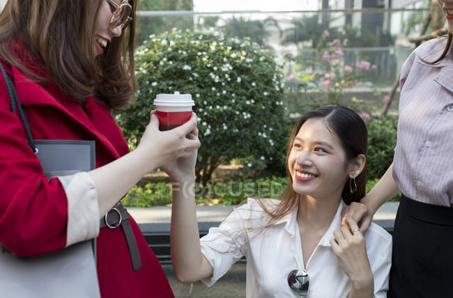 Улыбающаяся молодая женщина, дающая кофе подруге, сидя в городе — стоковое фото