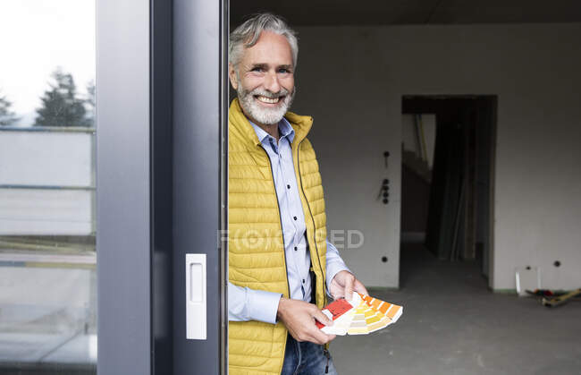 Sorridente architetto maschio che tiene campione di colore in casa ristrutturare visto attraverso la finestra — Foto stock