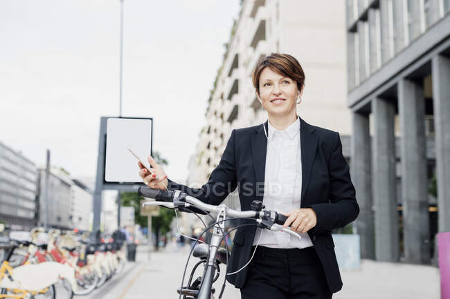 Female professional listening music while walking with bicycle on sidewalk in city - foto de stock