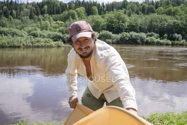 Sonriente hombre adulto medio instalando tienda de campaña mientras está de pie contra el lago en el bosque - foto de stock