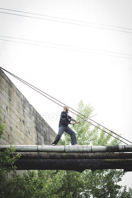 Maturo uomo tenendo spada posa mentre in piedi su vecchi tubi contro chiaro cielo — Foto stock