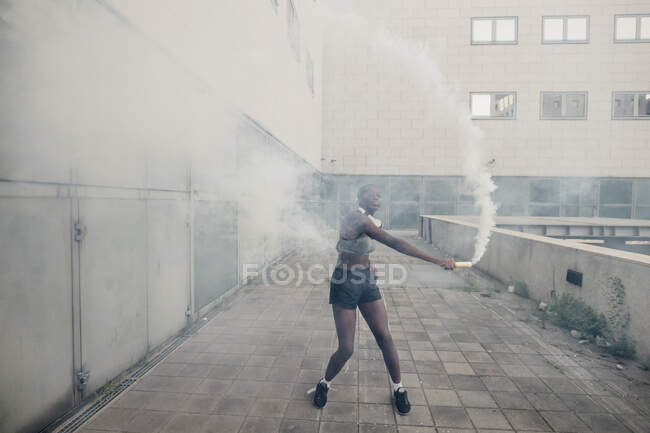 Молодая женщина играет с дымовой бомбой, стоя на пешеходной дорожке против здания в городе — стоковое фото