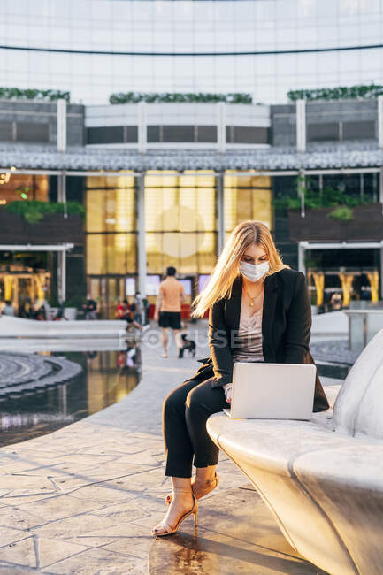 Geschäftsfrau trägt Maske mit Laptop, während sie auf Sitz gegen Gebäude sitzt — Stockfoto