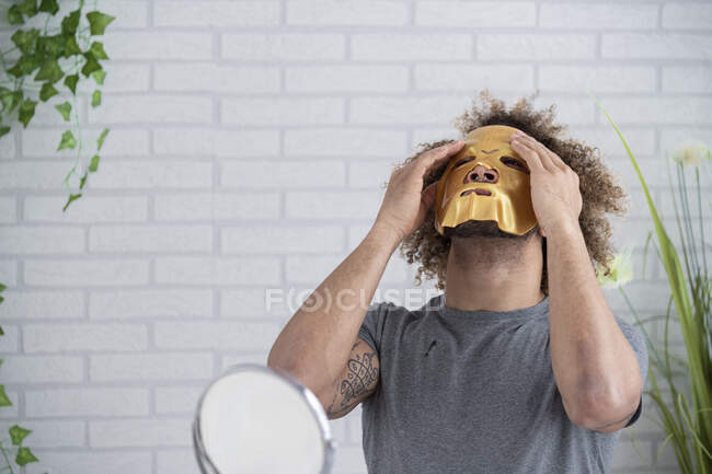 Jovem aplicando máscara facial enquanto sentado contra a parede em casa — Fotografia de Stock