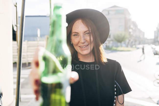 Gros plan d'une femme souriante tenant une bouteille de bière alors qu'elle se tenait en ville par une journée ensoleillée — Photo de stock