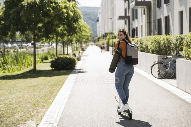 Donna sorridente guida scooter elettrico spinta su strada in città durante la giornata di sole — Foto stock