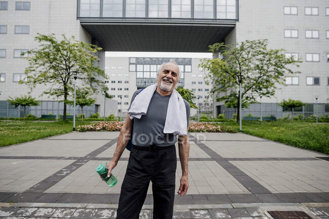 Щасливий старший, що тримає пляшку з водою, стоячи на стежці проти будівництва. — стокове фото