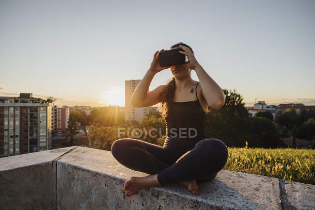 Sportliche Frau mit Vr-Brille während sie bei Sonnenuntergang auf Stützmauer in der Stadt sitzt — Stockfoto