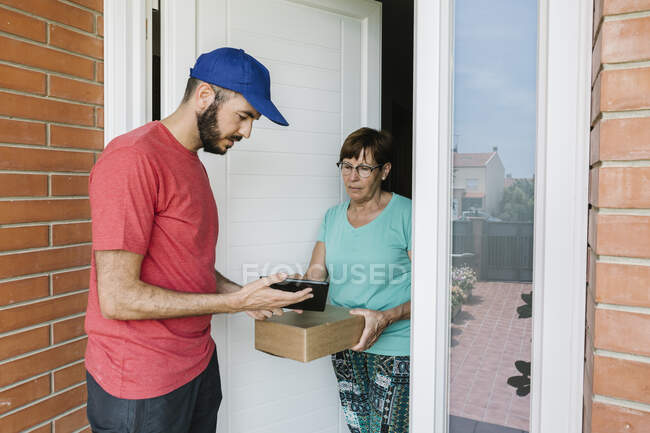 Trabajadora de correos mostrando tableta digital a una mujer mayor para su firma en la puerta - foto de stock
