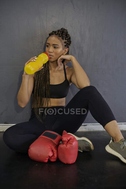 Atleta donna con capelli intrecciati che beve acqua seduta contro il muro in palestra — Foto stock