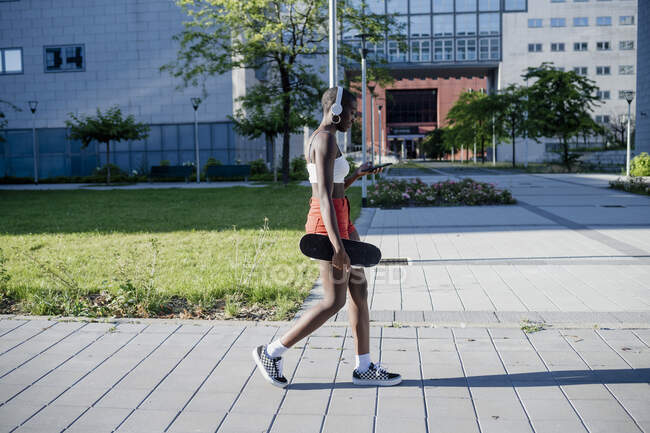 Young woman holding skateboard using smart phone while walking on footpath in city — Stock Photo