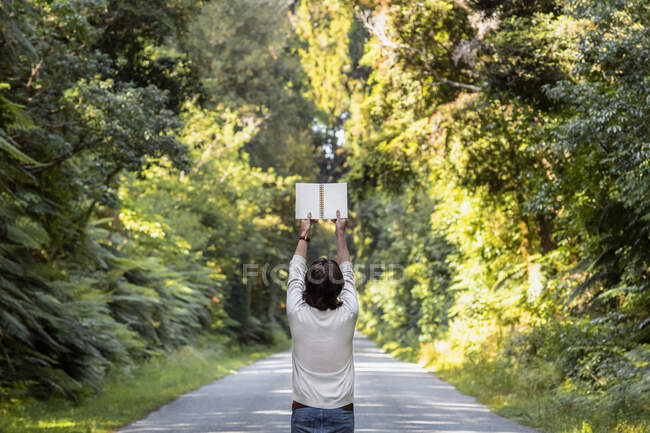 Young man holding blank diary while standing on road amidst trees in forest — Stock Photo