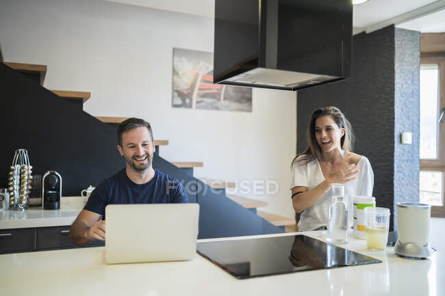 Smiling man working over laptop while woman preparing drink on kitchen island — Stock Photo