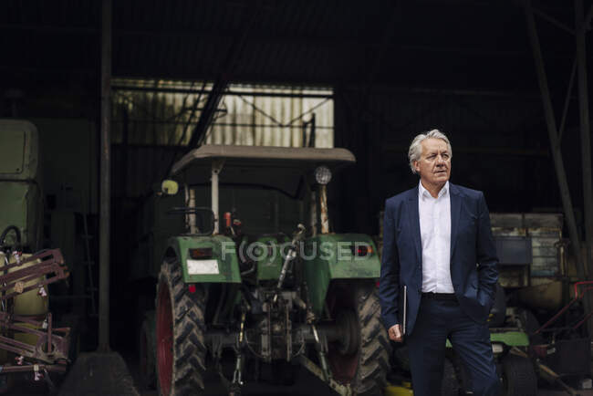 Empresário sênior segurando laptop em uma fazenda com trator no celeiro — Fotografia de Stock