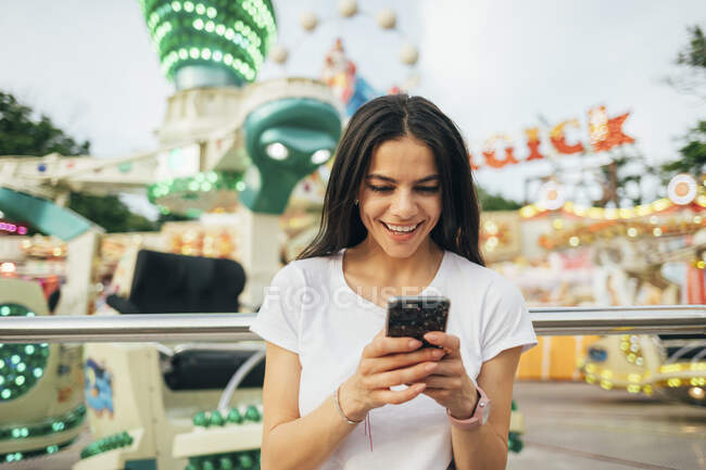 Mujer joven sonriente usando el teléfono inteligente mientras está de pie en el parque de atracciones - foto de stock