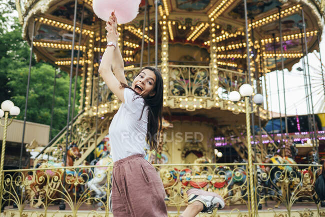 Mujer joven sosteniendo algodón de azúcar gritando mientras baila contra el carrusel en el parque de atracciones — Stock Photo
