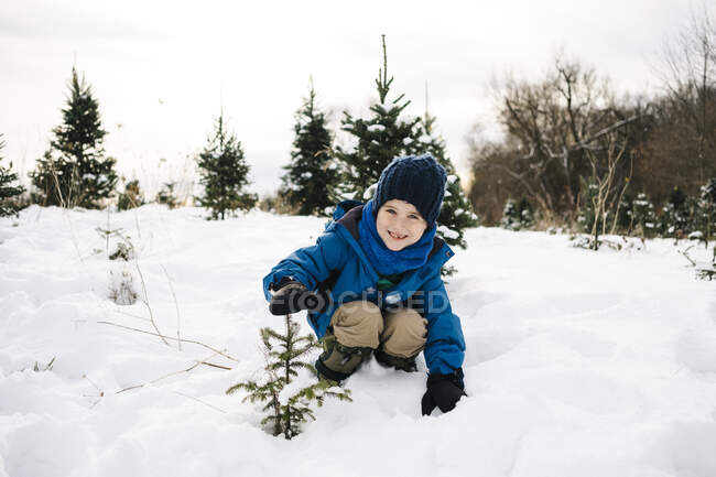 Усміхнений хлопчик показує дерево, присідаючись на вкритій снігом землі проти неба. — стокове фото
