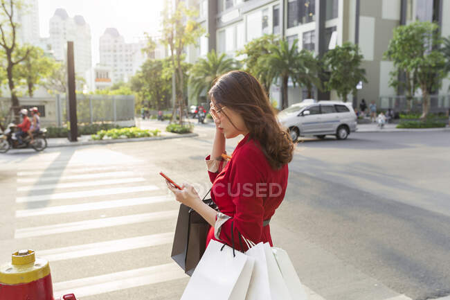 Mujer joven que lleva bolsas de compras usando el teléfono inteligente mientras está de pie en la calle - foto de stock