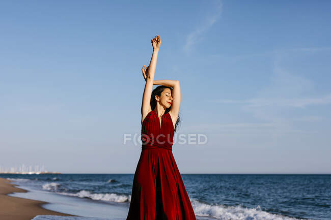 Delicate woman in red dress dancing at the sea, feeling the sun — Photo de stock