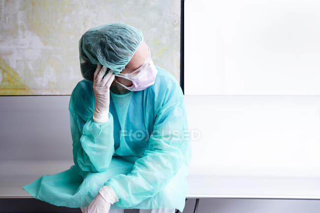Triste médecin portant des vêtements de travail protecteurs assis contre le mur à l'hôpital — Photo de stock