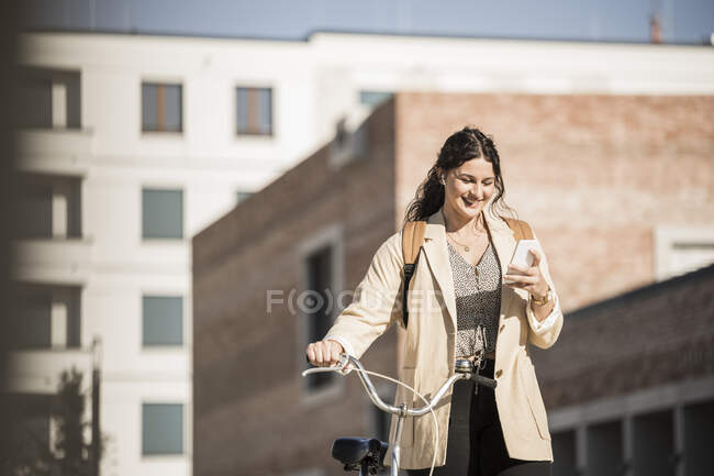 Lächelnde Pendlerin benutzt Handy, während sie mit dem Fahrrad gegen Gebäude in der Stadt läuft — Stockfoto
