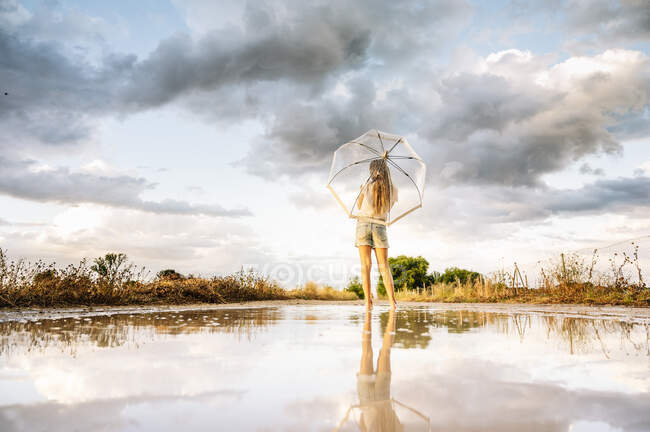 Chica pre-adolescente con paraguas de pie contra la reflexión en el charco - foto de stock