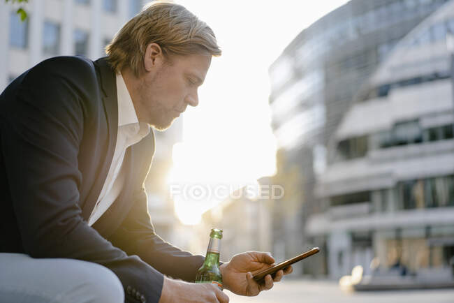 Homme d'affaires assis sur un banc de la ville au coucher du soleil avec une bouteille de bière et smartphone — Photo de stock