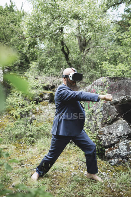 Empresário olhando através de simulador de realidade virtual praticando artes marciais contra árvores na floresta — Fotografia de Stock