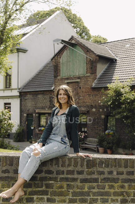 Portrait of smiling young woman sitting on a brick wall at a farmhouse — Stock Photo