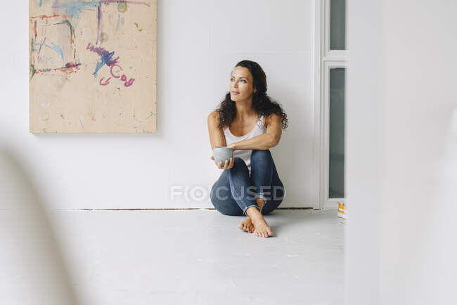Donna in possesso di tazza di caffè contemplando mentre seduto sul pavimento contro la parete in soppalco — Foto stock