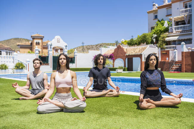Молодые многонациональные друзья медитируют во время занятий йогой на траве у бассейна на заднем дворе в солнечный день — стоковое фото
