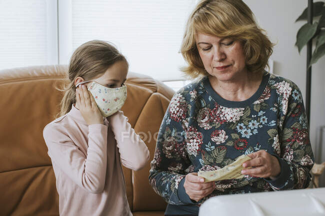 Fille portant un masque de protection fait maison tout en étant assis par grand-mère à la maison — Photo de stock