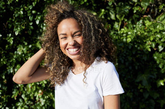 Fröhliche junge Frau steht an sonnigen Tagen mit der Hand im Haar gegen Pflanzen — Stockfoto