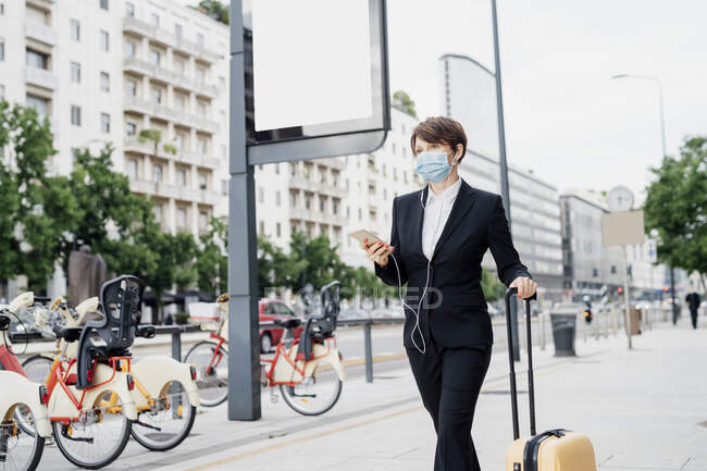 Businesswoman wearing mask listening music while walking with suitcase on sidewalk in city - foto de stock