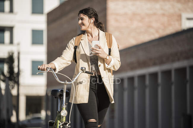 Усміхаючись, жінка дивиться убік, коли йде з велосипедом проти будинків у місті. — стокове фото