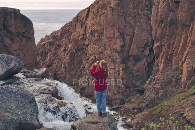 Mujer joven fotografiando cascada mientras está de pie en una montaña rocosa en Teriberka, Óblast de Murmansk, Rusia - foto de stock