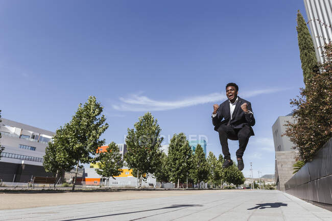Fröhlicher Geschäftsmann springt bei sonnigem Wetter auf Fußweg vor blauem Himmel in der Stadt — Stockfoto