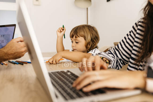 Cute daughter painting on paper while sitting with working parents in dining room — Stock Photo