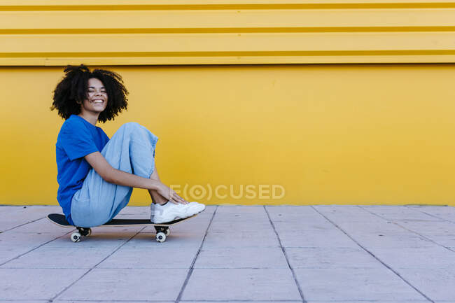 Giovane donna sorridente seduta sullo skateboard davanti al muro giallo — Foto stock