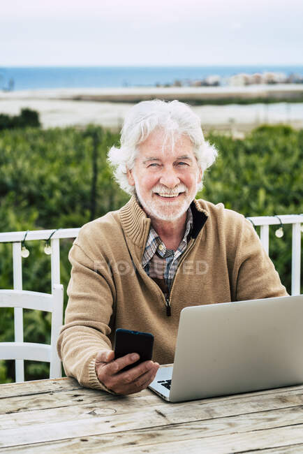 Sorrindo homem sênior segurando telefone inteligente enquanto sentado com laptop na mesa — Fotografia de Stock
