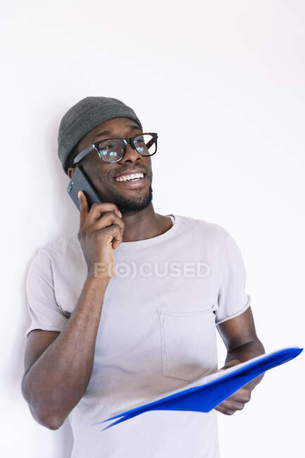 Sorrindo jovem empresário segurando arquivo e falando no telefone inteligente enquanto olha para o fundo branco — Fotografia de Stock