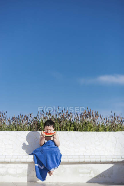 Carino ragazzo mangiare anguria fetta mentre seduto contro blu cielo — Foto stock