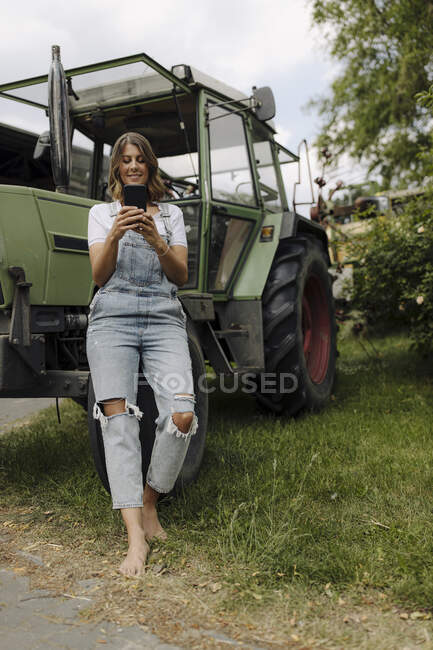 Jeune femme utilisant un téléphone portable à un tracteur dans la campagne — Photo de stock