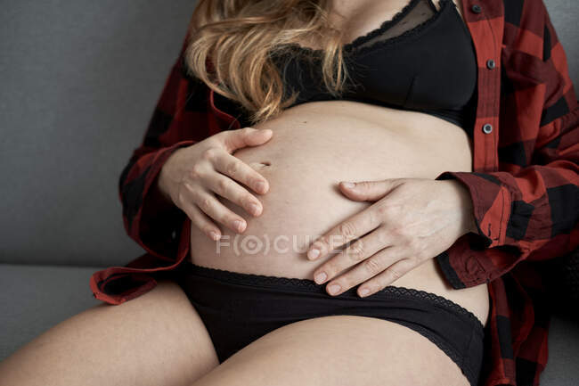 Primo piano della donna incinta con le mani sullo stomaco seduta sul divano a casa — Foto stock