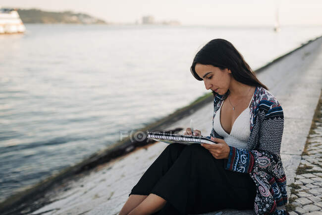 Mujer joven pintando sentada en la orilla del río - foto de stock